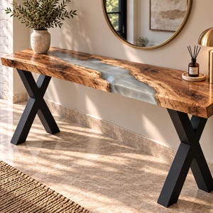 May include: A long console table featuring a natural live-edge wood slab top with a central translucent gray resin river inlay. The table is supported by two black metal X-shaped legs, providing a sturdy base for the elongated wooden surface.