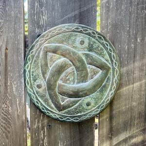 May include: A round, weathered green plaque with a Celtic knot design is mounted on a rustic wooden fence. The plaque features a detailed, three-part knot in the center, surrounded by an ornate border. The fence has vertical wooden planks with visible grain.