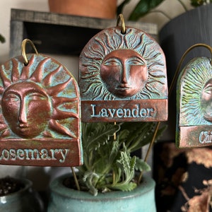 May include: Three rustic garden markers with a sun design and copper and turquoise paint. The markers are labeled "Rosemary", "Lavender", and "Chives".