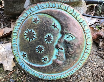 Puede incluir: Un paso de jardín circular de metal de color turquesa con un diseño de media luna y una cara. La luna está rodeada de cinco estrellas.