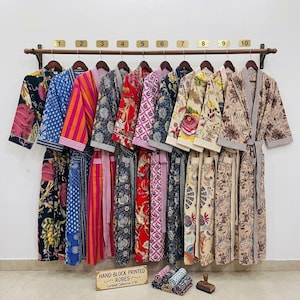 Könnte beinhalten: Eine Kollektion von zehn handbedruckten Roben in verschiedenen Farben und Mustern, die an einer Holzstange hängen. Die Roben weisen florale, geometrische und abstrakte Designs auf. Ein Schild zeigt "HAND-BLOCK PRINTED ROBES Curated Collection 1-10".