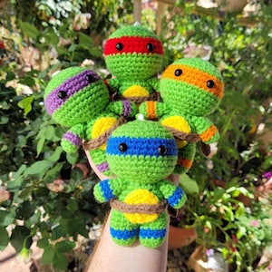 May include: A collection of four handmade, green crocheted turtle figures, each with a different colored mask: red, purple, orange, and blue. They have yellow bellies and are tied with brown yarn. The figures are displayed against a backdrop of green foliage.