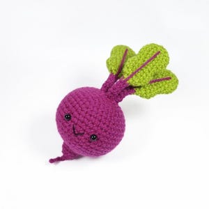 May include: A crocheted beet toy in shades of purple and green. The beet has a round body, a smiling face with black eyes, and green leaves with purple stems. The toy is handmade and sits against a white background.