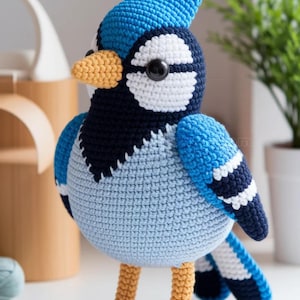 以下が含まれることがあります： 手作りのかぎ針編みのアオカケス。特徴的なとさか、黒と白の模様、黄色のくちばしと足が特徴です。鳥はさまざまな青色の糸で作られており、直立しており、その詳細なデザインを示しています。