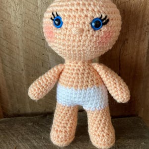 May include: A handmade crochet doll with a peach-colored body, blue button eyes, and rosy cheeks. The doll has a white diaper and is standing upright. The doll is made of yarn and is a soft, plush toy.