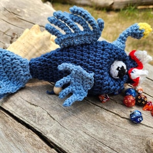 May include: A handmade blue crochet fish with detailed fins and a yellow-tipped antenna. The fish has a white eye with a black pupil and a red and white mouth. Assorted dice are scattered around the fish, suggesting a tabletop game theme.