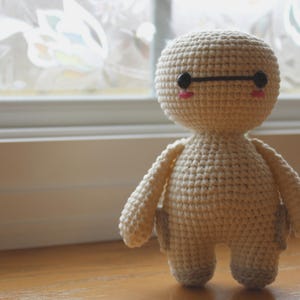 May include: A handmade, off-white crochet doll with a round head, small arms, and legs. The doll has black eyes, a black horizontal line above the eyes, and pink cheeks. It is standing on a wooden surface near a window.