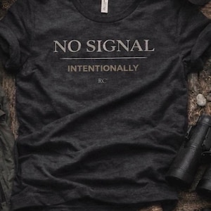 May include: Dark gray t-shirt with the text "NO SIGNAL" above "INTENTIONALLY". The shirt has a crew neck and short sleeves. A black cap and binoculars are visible in the background.