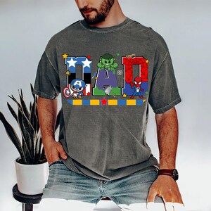 Op de afbeelding: Grijs t-shirt met het woord "DAD" in kleurrijke letters, elk versierd met een ander superheldenpersonage. De letter "D" heeft een blauw-wit sterrenpatroon, de "A" heeft de Hulk en de laatste "D" heeft Spider-Man.