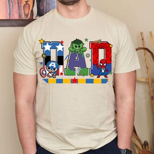 Op de afbeelding: Beige t-shirt met het woord "DAD" in een kleurrijk ontwerp met Captain America, Hulk en Spider-Man. De letters zijn versierd met sterren en superheldenafbeeldingen, waardoor het een leuke, casual tee is.