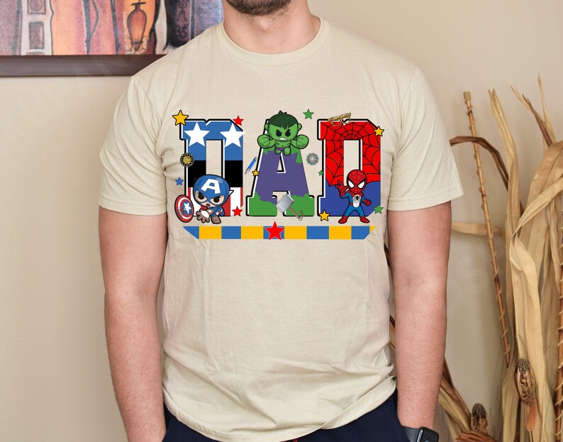 Op de afbeelding: Beige t-shirt met het woord "DAD" in een kleurrijk ontwerp met Captain America, Hulk en Spider-Man. De letters zijn versierd met sterren en superheldenafbeeldingen, waardoor het een leuke, casual tee is.