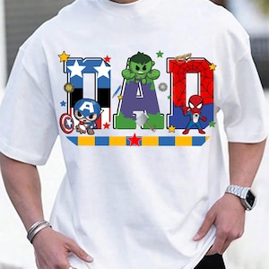 Op de afbeelding: Wit T-shirt met het woord "DAD" in een kleurrijk superheldenontwerp. De letters zijn versierd met sterren en superheldenpersonages. Een blauw-gele en rode streep loopt over de onderkant.