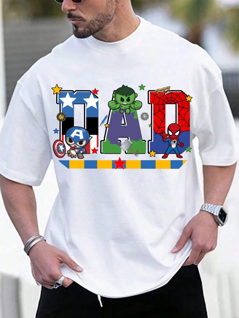 Op de afbeelding: Wit T-shirt met het woord "DAD" in een kleurrijk superheldenontwerp. De letters zijn versierd met sterren en superheldenpersonages. Een blauw-gele en rode streep loopt over de onderkant.
