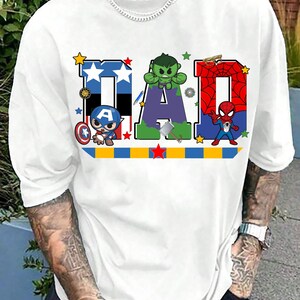 Op de afbeelding: Wit T-shirt met het woord "DAD" in kleurrijke blokletters. Elke letter is ontworpen met een superheldenthema, met Captain America, Hulk en Spider-Man. Een blauw, geel en rode streep loopt over de onderkant.