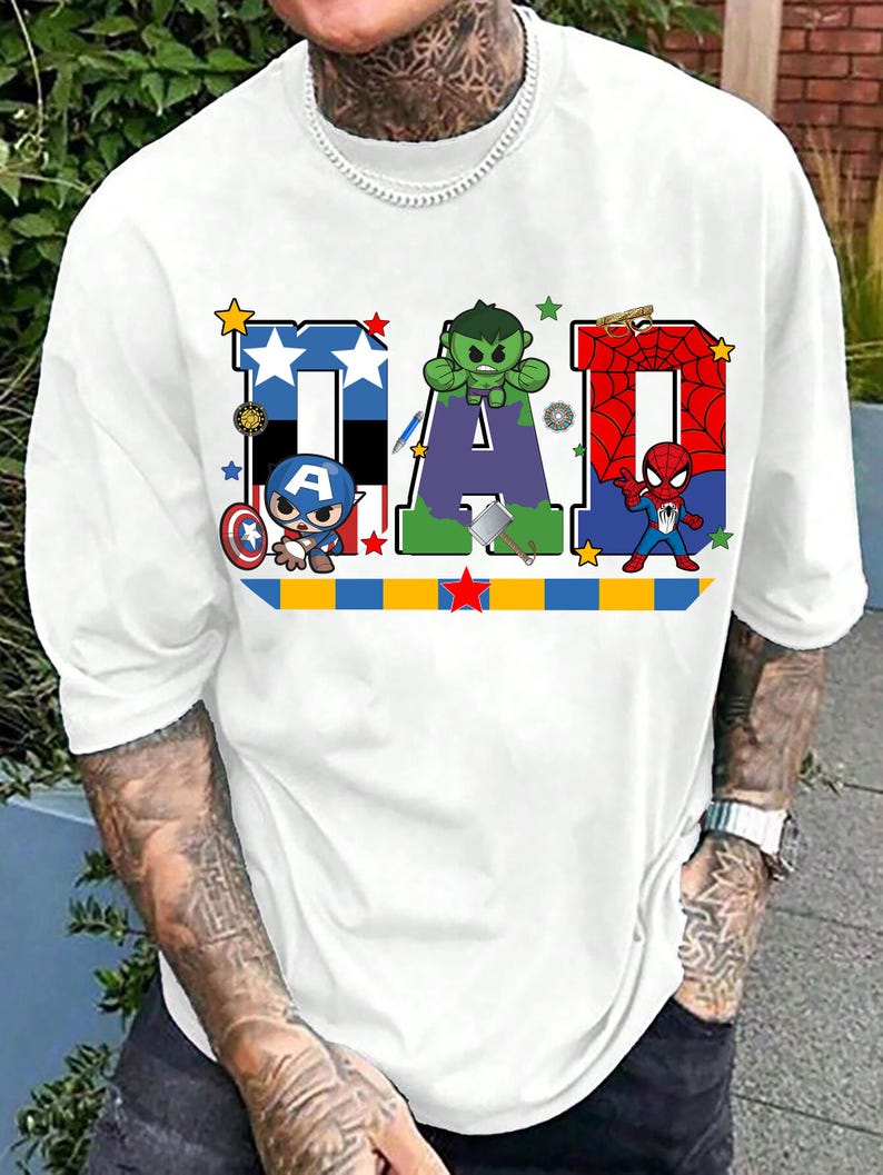 Op de afbeelding: Wit T-shirt met het woord "DAD" in kleurrijke blokletters. Elke letter is ontworpen met een superheldenthema, met Captain America, Hulk en Spider-Man. Een blauw, geel en rode streep loopt over de onderkant.