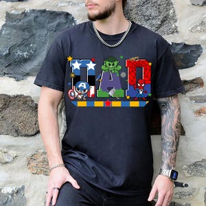 Op de afbeelding: Zwart t-shirt met het woord "DAD" in een kleurrijk ontwerp met superheldenpersonages. De letter "D" is blauw met witte sterren, "A" is groen met Hulk en "D" is rood met Spider-Man. Er is ook een Captain America-afbeelding.