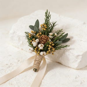 Peut inclure: Boutonni&egrave;re compos&eacute;e d'un petit c&ocirc;ne de pin, de boutons blancs ressemblant &agrave; du coton, de baies jaunes et de feuillage vert. Les tiges sont envelopp&eacute;es de ficelle et attach&eacute;es avec un ruban cr&egrave;me. Id&eacute;al pour les mariages.