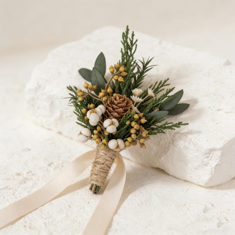 Peut inclure: Boutonni&egrave;re compos&eacute;e d'un petit c&ocirc;ne de pin, de boutons blancs ressemblant &agrave; du coton, de baies jaunes et de feuillage vert. Les tiges sont envelopp&eacute;es de ficelle et attach&eacute;es avec un ruban cr&egrave;me. Id&eacute;al pour les mariages.