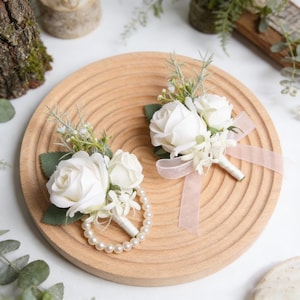 May include: Two white rose boutonnieres with greenery and a pearl bracelet corsage on a wooden tray. One boutonniere has a pink ribbon. The flowers are arranged with green leaves and small white flowers.