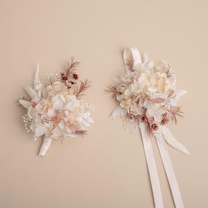 K&ouml;nnte beinhalten: Zwei Str&auml;u&szlig;e aus Trockenblumen in Creme-, Pfirsich- und Braunt&ouml;nen. Ein Strau&szlig; ist mit einem cremefarbenen Band gebunden, der andere mit einem cremefarbenen Stoff umwickelt. Die Blumen umfassen Hortensien und andere zarte Bl&uuml;ten.