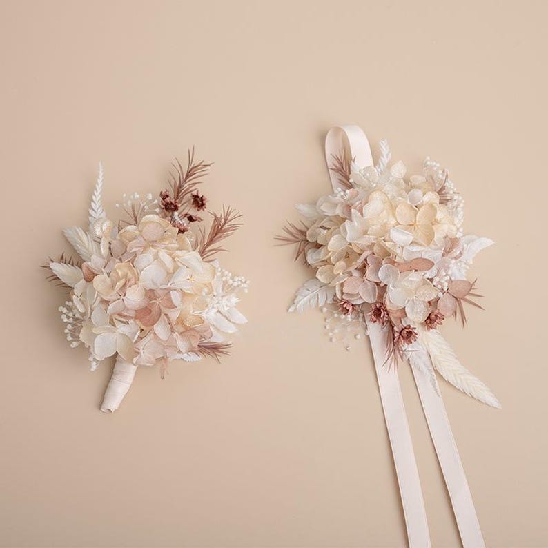 K&ouml;nnte beinhalten: Zwei Str&auml;u&szlig;e aus Trockenblumen in Creme-, Pfirsich- und Braunt&ouml;nen. Ein Strau&szlig; ist mit einem cremefarbenen Band gebunden, der andere mit einem cremefarbenen Stoff umwickelt. Die Blumen umfassen Hortensien und andere zarte Bl&uuml;ten.