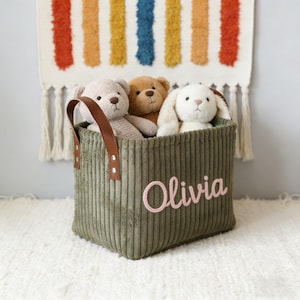 May include: A green corduroy storage basket with the name "Olivia" embroidered in pink. The basket contains a beige teddy bear, a brown teddy bear, and a white bunny. The basket has brown leather handles.