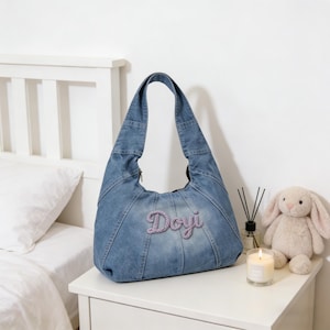 May include: A light blue denim shoulder bag with the name "Doyi" embroidered in a cursive, light purple font. The bag has a curved shape and a zipper closure. The bag is sitting on a white nightstand next to a stuffed bunny and a candle.