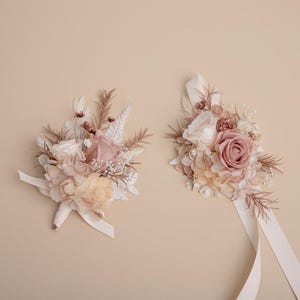 K&ouml;nnte beinhalten: Zwei Blumenarrangements mit Rosen und anderen Bl&uuml;ten in Rosa-, Creme- und Beiget&ouml;nen. Eines ist ein Strau&szlig; mit Schleife, das andere ein Gesteck mit langen B&auml;ndern. Der Hintergrund ist einfarbig beige.