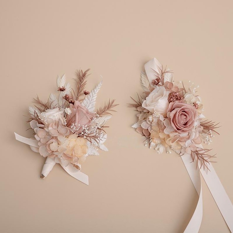 K&ouml;nnte beinhalten: Zwei Blumenarrangements mit Rosen und anderen Bl&uuml;ten in Rosa-, Creme- und Beiget&ouml;nen. Eines ist ein Strau&szlig; mit Schleife, das andere ein Gesteck mit langen B&auml;ndern. Der Hintergrund ist einfarbig beige.