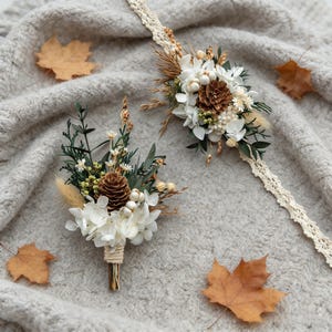 May include: A boutonniere and a wrist corsage featuring dried white flowers, pinecones, and greenery. The boutonniere is wrapped with twine. The wrist corsage is attached to a cream-colored lace band. The items are arranged on a gray knitted surface with scattered fall leaves.