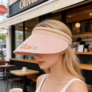 May include: A pink straw visor with a beige and brown striped band. The visor has the name "Olivia" embroidered in colorful letters. The visor is designed to shield the eyes from the sun.