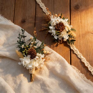 Peut inclure: Deux compositions florales rustiques. L'une est une boutonni&egrave;re avec une pomme de pin, des fleurs blanches et de la verdure, attach&eacute;e avec de la ficelle. L'autre est un bracelet de corsage avec des &eacute;l&eacute;ments similaires, dont une bande de dentelle. Sur une surface en bois.
