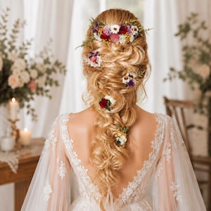 May include: A long, braided blonde hairstyle adorned with multiple small clusters of dried flowers in shades of red, white, purple, and yellow. The hair is styled in a loose, textured braid, with a larger floral arrangement placed at the crown.