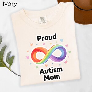 Peut inclure: T-shirt ivoire avec les mots "Proud Autism Mom" en noir. Un symbole infini arc-en-ciel est centré entre les mots, avec de petits cœurs et étoiles autour du motif. Le t-shirt est fait d'une matière douce.