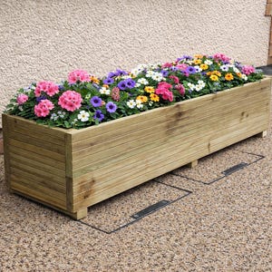 May include: A long, rectangular wooden planter filled with a vibrant mix of flowers. The flowers are in shades of pink, purple, yellow, and white. The planter is made of light-coloured wood and is on a stone surface.