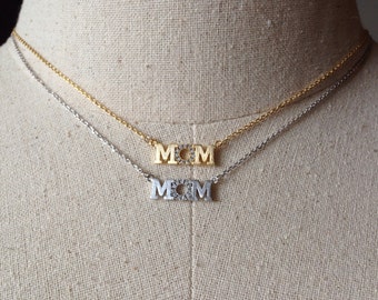 Puede incluir: Dos collares de oro y plata con la palabra "MOM" escrita en letras de bloque con pequeños diamantes de imitación transparentes alrededor de cada letra.