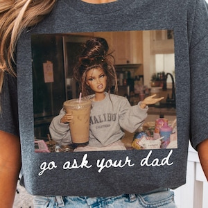 Op de afbeelding: Donkergrijs t-shirt met een afbeelding van een pop die een koffiekopje vasthoudt, met een grijze sweatshirt met de tekst "Malibu California". Het shirt heeft de tekst "go ask your dad" in witte letters.