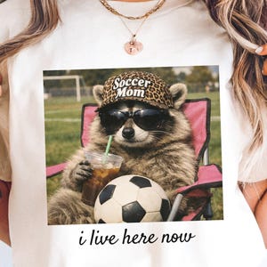 Puede incluir: Camiseta blanca con un mapache con gafas de sol y un sombrero con estampado de leopardo que dice "Soccer Mom". El mapache sostiene una bebida y est&aacute; sentado en una silla rosa junto a un bal&oacute;n de f&uacute;tbol. El texto "i live here now" est&aacute; debajo.