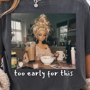 Peut inclure: Un t-shirt gris anthracite avec une impression graphique repr&eacute;sentant une poup&eacute;e aux cheveux blonds en chignon d&eacute;coiff&eacute;, assise &agrave; une table de cuisine avec un caf&eacute; et un bol de c&eacute;r&eacute;ales. Le texte en dessous indique "too early for this".
