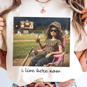 Puede incluir: Camiseta color crema con una imagen impresa de una muñeca con gafas de sol, ropa con estampado de leopardo y un sombrero, sentada en una silla rosa en un campo de béisbol. El texto "i live here now" está impreso debajo.