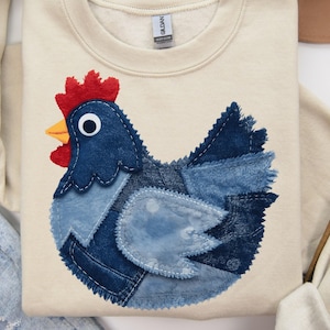 Peut inclure: Un sweat-shirt crème avec un motif de poulet en patchwork de denim. Le poulet a une crête rouge, un bec jaune et un grand œil. Le motif est fait de différentes nuances de denim bleu.