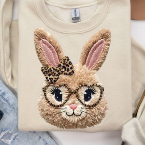 May include: Cream-colored sweatshirt featuring a detailed bunny face design. The bunny has fluffy brown fur, pink inner ears, and wears heart-shaped glasses and a leopard print bow. A cute and festive design.