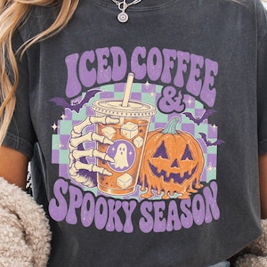 May include: A charcoal gray t-shirt featuring a graphic print of a skeleton hand holding an iced coffee cup next to a melting orange pumpkin. The text reads ICED COFFEE & SPOOKY SEASON, surrounded by purple bats and a light green and purple checkerboard.