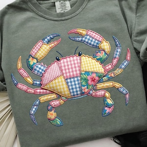 May include: Sage green long-sleeve shirt featuring a colorful crab design. The crab is made of patchwork fabric in pink, yellow, blue, and floral patterns. The shirt has a crew neck and a tag that says "Comfort Colors".