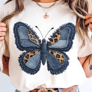 以下が含まれることがあります： 大きな蝶のデザインが施されたクリーム色のTシャツ。蝶は、縁がほつれたデニムブルーの羽と、ヒョウ柄のアクセントが付いています。シャツは丸いネックラインと半袖です。