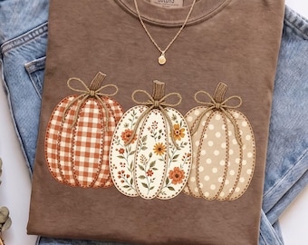 May include: A brown short-sleeve t-shirt with a Comfort Colors label at the neck, featuring a graphic of three pumpkins with twine bows. The left pumpkin has an orange gingham pattern, the center has a floral print, and the right has white polka dots.