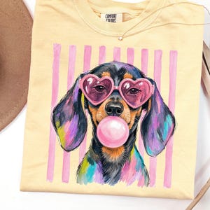 Puede incluir: Una camiseta de color amarillo pálido con el gráfico de un perro salchicha negro y fuego que lleva gafas de sol rosas en forma de corazón y hace una burbuja rosa. El perro está sobre un fondo de rayas verticales rosas. Tiene etiqueta Comfort Colors.