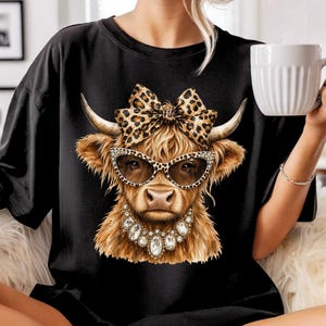 May include: A black t-shirt featuring a graphic print of a brown highland cow wearing leopard print glasses, a matching leopard print bow on its head, and a large, multi-faceted clear crystal statement necklace around its neck.