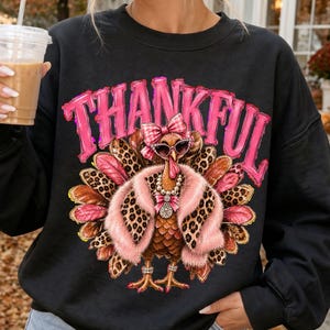Könnte beinhalten: Ein schwarzes Langarm-Sweatshirt mit einer Grafik eines Truthahns, der eine rosa Pelzstola, eine Perlenkette, eine Sonnenbrille und eine karierte Schleife trägt. Das Wort THANKFUL ist in großen, abgenutzten rosa Buchstaben darüber gedruckt.