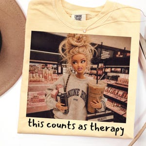 Op de afbeelding: Een lichtgeel t-shirt met een grafische print van een pop met blond haar in een rommelige knot, gekleed in een grijze sweater met de tekst NINE BINE SPORT, met winkeltassen en een koffiebeker in een winkel. De tekst this counts as therapy staat onderaan.